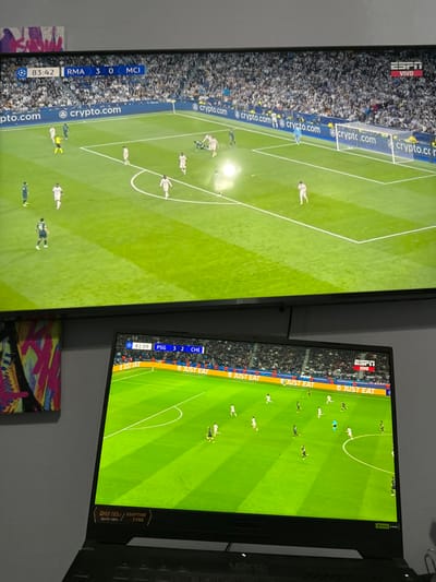This image depicts an indoor setting in the city of El Hueco, Argentina, where two electronic displays are simultaneously showing live soccer matches.
The upper display is a large, wall-mounted television screen. It features a soccer match between Real Madrid (RMA) and Manchester City (MCI), with the score reading RMA 3 - 0 MCI at 83 minutes and 42 seconds. The broadcast is identified as ESPN VIVO, and "crypto.com" is visible on the stadium's perimeter advertising boards. The field shows players in white and blue uniforms on a green pitch, with several players clustered near a goal and a light blue-clad goalkeeper. A prominent bright reflection is visible on the screen's surface. The stadium background is densely populated with spectators.
Below the television, a black laptop is open, displaying a different soccer match. The laptop's screen shows a game between Paris Saint-Germain (PSG) and Chelsea (CHE), with the score PSG 3 - 2 CHE at 81 minutes and 9 seconds. This match is also broadcast by ESPN VIVO, and "JUST EAT" is visible on the stadium's perimeter advertisements. Players in dark and light jerseys are distributed across the green field, with spectators visible in the background stadium seating. The laptop's bezel partially reveals "ASUS TUF" branding along with "QHD 165Hz | ADAPTIVE SYNC."
The immediate environment includes a plain, light-colored wall. To the left of the laptop, an abstract artwork is mounted, featuring vivid pink, yellow, and blue geometric or chaotic shapes against a darker background. No individuals are directly visible in the image.
