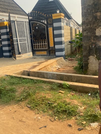 This media file captures the entrance of a residential property in Igbesa, Nigeria, presented with a vertical orientation. The central feature is a house with a striking black and gold wrought iron gate.
The house displays a modern architectural style. Its exterior walls feature alternating horizontal stripes of black and light gray, mirroring the design on the gate pillars. The gate itself is black with prominent gold accents, showcasing intricate scrollwork and geometric patterns. It includes both a larger main gate and what appears to be a smaller pedestrian gate within it. The roof of the house, partially visible, is dark-colored, likely tiled.
To the right of the house and gate, a light-colored, possibly concrete or paved, pathway or driveway extends forward. This pathway is bordered by a concrete drainage channel running parallel to it. Beyond the channel, the ground is unpaved, composed of reddish-brown soil, interspersed with patches of green grass. There is some scattered debris and small pieces of litter visible on the unpaved ground.
The lighting indicates it is daytime, with bright sunlight illuminating the scene. No individuals or specific activities are visible, presenting a static view of the property's exterior. No distinct text is visible on any part of the house, gate, or surrounding environment. The setting suggests a residential area, possibly on the outskirts or a developing part of a town, characterized by both constructed elements and natural, unmanicured ground.
