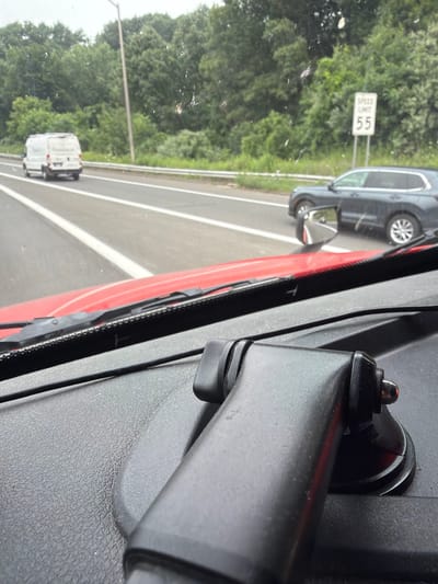 An eye-level, slightly high-angle, medium shot captures a view from inside a vehicle traveling on a multi-lane road. In the foreground, the dashboard of the vehicle is visible, with a black textured surface and a red dashboard accent. A black dashboard mount for a camera or electronic device is prominently featured in the lower right and center of the frame. Beyond the dashboard, the road ahead is visible with white lane markings.
In the mid-ground, a white van is traveling in the left lane, and further to the right, a dark-colored SUV is in the right lane. Both vehicles are moving away from the viewer. A speed limit sign indicating "55" is visible on the right side of the road, surrounded by lush green trees that line the highway. The sky appears overcast. The context of Danbury, United States of America, is not directly discernible from the visual elements of the image.