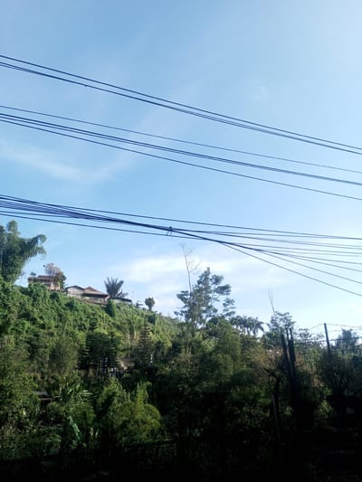 The image shows a view of a verdant, tree-covered hillside with a few buildings visible at the top, situated in Phou Khoun, Laos. Several utility wires stretch across the upper portion of the frame against a light blue sky with scattered clouds. The foliage is dense and varied, suggesting a tropical or subtropical environment. A section of a fence is visible on the lower right side. The image gives the impression of a rural or semi-rural setting within Phou Khoun. There are no people visible in the picture.