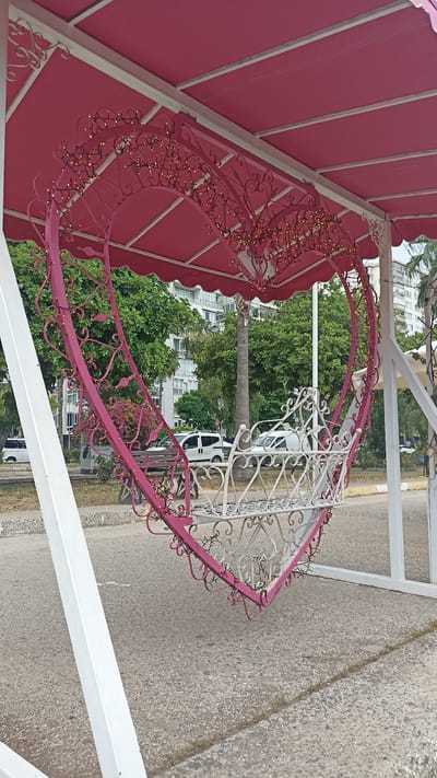 This is a photograph taken in Mersin, Turkey, of a decorative heart-shaped swing. The swing is suspended from a pink awning supported by white posts. The heart-shaped frame of the swing is adorned with small, yellow lights. Inside the heart, there is a white, ornate bench. In the background, there are buildings, trees, and parked vehicles, indicating an urban setting. The ground is paved with asphalt. The image appears to be taken during the daytime under overcast skies.