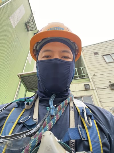 In Geoje-si, South Korea, a construction worker is captured in a selfie, standing outside a light green industrial building. The worker is wearing a dark blue long-sleeved shirt, a dark blue face mask, and an orange hard hat with "SAMSUNG" visible on its front. A safety harness, primarily blue with yellow accents and a red and blue rope attached, is worn over the shirt. Safety glasses are resting on the harness. The background features the exterior of the industrial building, with a white-patterned section and a metal grate structure on the green wall. Another building, primarily light beige, is visible to the right, with windows and vents. The sky is overcast and white.