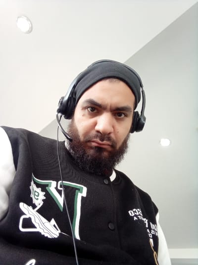 A man is featured in a close-up, low-angle shot, looking directly at the camera with a stern or serious expression. He has a full dark beard and is wearing a black beanie or headwrap that covers his hair. Over the beanie, he wears a black headset with a microphone extending towards his mouth.
He is dressed in a black varsity-style jacket with white sleeves. On the left side of his jacket, there are prominent white and green graphics, including a large letter "V" outlined in green, a white graphic resembling a shoe or boot, and a cog-like or star shape. On the right side of the jacket, partially visible and appearing inverted or reversed due to the angle, are white letters and numbers that read "03D A TO..." and "4 213 D".
The background consists of a plain white ceiling with two bright, circular recessed light fixtures visible. The lighting is bright, suggesting an indoor setting during the daytime. The perspective of the photograph is from below, looking up at the man. The location context for this image is Parroquia Guatire, Venezuela.