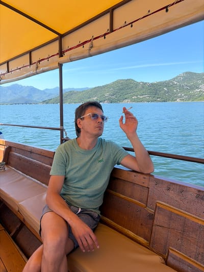 A single adult male is seated on a wooden boat, observed from the chest up on the right side of the frame. He wears a light green short-sleeved t-shirt, patterned shorts, and clear-framed sunglasses. His left hand, bent at the elbow and resting on the boat's wooden railing, holds a lit cigarette. His gaze is directed towards the left (viewer's left), across a large body of water. The boat features wooden paneling, light-colored upholstered benches, and a yellow canvas canopy supported by dark metal poles. Below the canopy, a white fabric lining with red cording and clear plastic roll-up panels is visible. To the immediate left of the man, a small container labeled "TIPS FOR CAPTA" is situated on a small shelf.
The background depicts a vast, calm body of blue-green water under a clear, bright blue sky. Across the water, a long, low bridge with multiple spans is visible in the middle distance. Beyond the water and bridge, a series of green, tree-covered mountains rise under the clear sky. The scene takes place in the vicinity of Vranjina, Montenegro.