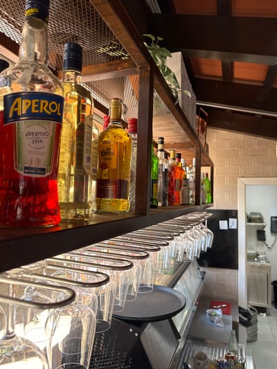 This is a close-up, low-angle shot of a shelf stocked with various bottles of alcohol, with a rack of clear glasses to the right. The bottles, predominantly in amber, red, and green hues, are arranged on metal shelves with a wire mesh backing. In the foreground, a bottle labeled "APEROL" with Italian text is prominently displayed. Behind it, other bottles, including one identified as "Jose Cuervo Especial" tequila and "VERMO" bottles, are visible. The setting appears to be a bar or restaurant, with a hint of wooden paneling and textured walls in the background. The lighting suggests it could be daytime or evening with artificial lighting. To the right, a series of clean, empty wine or cocktail glasses are neatly arranged on a metal rack, catching the light. The overall impression is one of an organized bar ready for service. The visible text includes brand names and origin details such as "APEROL RICETTA ORIGINALE APERITIVO - DAL 1919 PADOVA-ITALIA," "Jose Cuervo Especial," and "BLUE AGAVE REPOSADO TEQUILA."