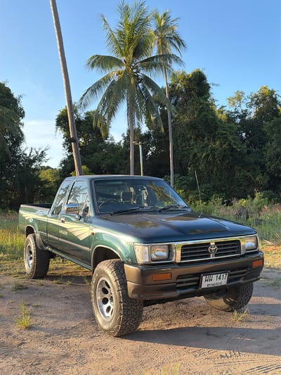 This media file is a photograph of a dark green Toyota Hilux pickup truck parked on a dirt road. The truck is oriented vertically in the frame. It has a chrome grill with the Toyota logo, square headlights, and a visible license plate. The license plate reads "ผผ 1417" and has Thai text below it. The truck appears to have off-road tires.

The setting is outdoors, with lush green trees and palm trees visible in the background. The sky is a clear, bright blue, suggesting it is daytime and the weather is fair. The sun is casting shadows, indicating it is likely late afternoon or early morning. The ground is dry and dusty, typical of a rural or unpaved road.

There are no people visible in the image. The focus is solely on the truck and its surroundings. The overall mood is peaceful and serene, capturing a moment in a tropical or subtropical environment. The location context is Baan Taling Ngam, Thailand, which aligns with the presence of palm trees and the style of the license plate.