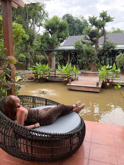 In Mae Rim, Thailand, a woman with auburn hair, wearing a leopard print jumpsuit and beige sneakers, reclines on a dark woven circular lounge chair. The chair rests on a reddish-brown tiled patio overlooking a murky pond with a small fountain. Lush green foliage and trees surround the pond, with a wooden deck and walkway extending over the water. In the background, a dark-tiled roof of a building is visible amidst the trees. The sky is overcast.