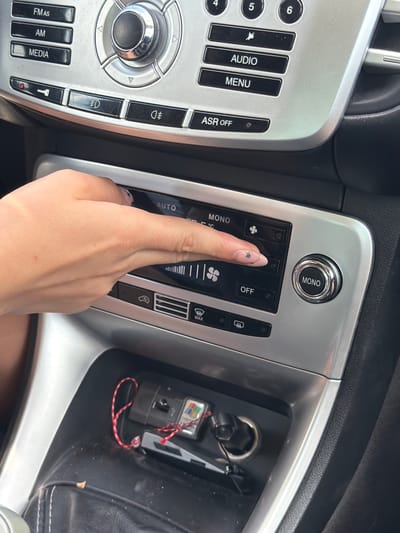 The image shows the interior of a car's dashboard, focusing on the climate control panel and some surrounding buttons. A person's hand is visible, with painted nails, touching the climate control display. The climate control panel features buttons labeled "AUTO" and "MONO," along with icons representing fan speed and temperature. There are also other buttons on the dashboard, including "FM/AS," "AM," "MEDIA," "AUDIO," "MENU," and "ASR OFF." Below the climate control panel, there is a storage compartment containing car keys and a USB drive. The dashboard and surrounding car interior appear to be made of silver-colored plastic. The scene is well-lit, suggesting it was taken during the day, possibly inside a vehicle parked in an urban environment. The presence of a Windows logo on the USB drive suggests a modern technological context. The overall impression is one of typical car interior functionality. This image is not located in Aleksandrovo, Bulgaria.
