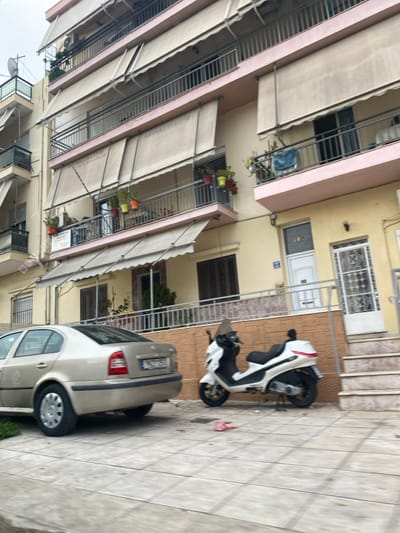 The image captures a residential scene in Koridallos, Greece, likely during the daytime under an overcast sky. The photograph is oriented vertically, showing a street or driveway flanked by buildings.
On the left, a multi-story apartment building features numerous balconies, each equipped with a light-colored retractable awning that is extended. Some balconies display potted plants with colorful flowers, while one has a blue item, possibly laundry, hanging. Air conditioning units are visible on some balconies. The facade of this building appears to be a light, possibly peach or pinkish hue.
To the right, a yellow-colored building is visible, featuring various windows and doors. One white entrance door has the number "215" affixed to the wall next to it. Security grilles protect some windows and doors, and a set of exterior stairs leads up to an entrance.
Parked on the tiled pavement in the foreground is a light-colored sedan, possibly a Skoda Octavia, facing away from the viewer. Behind it, closer to the yellow building, two white and black scooters are parked side-by-side. The pavement itself is made of large, square tiles. A small tree or bush is visible in the background between the buildings.
Visible text includes the number "215" next to a door and partially visible lettering on one of the awnings on the left building, which appears to say "SOLOMONIS". The scene is calm, depicting an ordinary moment in a residential neighborhood.