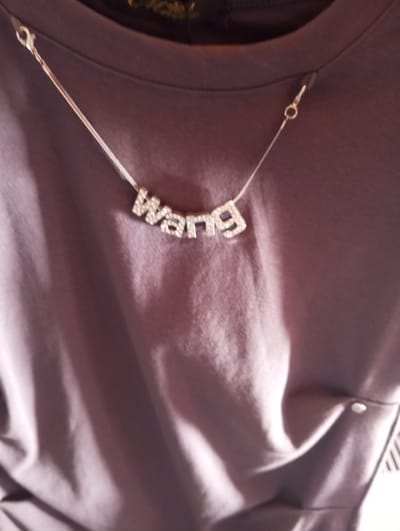The image shows a close-up of a purple shirt with a delicate silver necklace. The necklace features the word "wang" spelled out in blocky, rhinestone-encrusted letters. The necklace is attached to the shirt with small clasps on either side, near the collar. The shirt appears to be made of a soft, possibly silk or satin-like material, and is draped loosely. There is a slight sheen on the fabric, suggesting it is catching the light. The background is out of focus and consists of more of the purple fabric. The overall impression is one of subtle luxury and branding. There is no indication of the city of Velykyi Sukhodil, Ukraine, in this image.