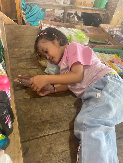 The image shows a young girl lying on a wooden surface, likely a stall or counter, in what appears to be a market setting in Ta', Indonesia. The girl is wearing a pink t-shirt and light blue pants. She is holding a small, rectangular object in her hands and looking down at it. Her hair is dark and she has pink barrettes in it. The background shows shelves with various products, including what look like toiletries and food items. There are also some cardboard boxes and a person's legs in a turquoise garment in the upper left corner of the image, suggesting a busy market environment. The wooden surface is worn and has visible grain and some imperfections.