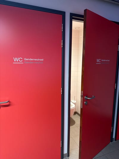 The image shows two red doors indicating gender-neutral restrooms in Rotterdam, Netherlands. The door on the left is closed, while the door on the right is ajar, revealing a glimpse of a toilet and a toilet brush within a tiled stall. Both doors are marked with "WC" and the text "Genderneutraal" and "Gender-neutral" in white lettering. The doors have silver door handles and appear to be part of a public facility.
