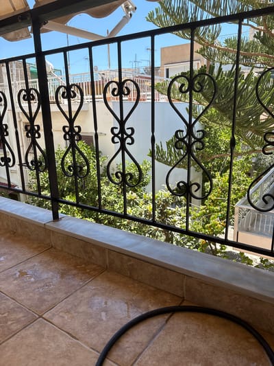 The image shows a view from a balcony in Ilion, Greece, looking out through an ornate black metal railing. The railing features decorative heart shapes and swirls. Beyond the railing, a dense collection of green trees and foliage is visible, some with small yellow fruits. In the background, white and tan buildings with visible antennas and air conditioning units suggest an urban or suburban setting. The sky is a clear blue. The foreground of the balcony is tiled in a sandy brown color, with some water patches and a black hose visible on the floor. A light-colored awning is stretched overhead, partially obscuring the sky.