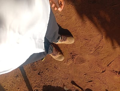 The image shows a person standing on reddish-brown dirt. The person is wearing dark pants and light brown sneakers with brown laces. Part of a white garment, possibly a lab coat or uniform, is visible on the left side of the frame, obscuring the rest of the person's body. The dirt ground has several shoe prints imprinted on it, indicating recent activity. Sunlight casts distinct shadows on the ground, suggesting it is daytime. The lighting and dry appearance of the dirt hint at a sunny, possibly warm, environment. The scene appears to be outdoors, in a location that is not paved. The visual cues of dirt, shadows, and footprints are consistent with a rural or less developed area. There is no visible text in the image. The context provided, Ukukwa, Nigeria, aligns with the visual characteristics of the scene, suggesting a possible outdoor gathering, work, or leisure activity in that location.
