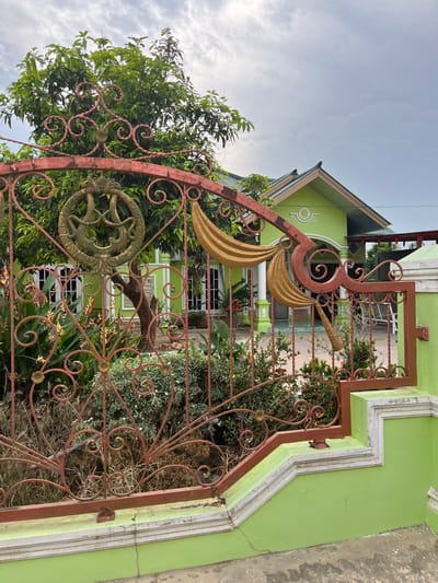 The image depicts the front of a bright green house, viewed from behind a red metal gate. The gate has intricate scrollwork and a circular emblem featuring a stylized, possibly mythical, creature. In front of the house, there is lush green foliage, including trees and flowering plants. The house itself has a green roof and white trim around the windows. A portion of a concrete walkway or patio is visible in the foreground. The sky is overcast. The location is identified as Tanahterban, Indonesia.