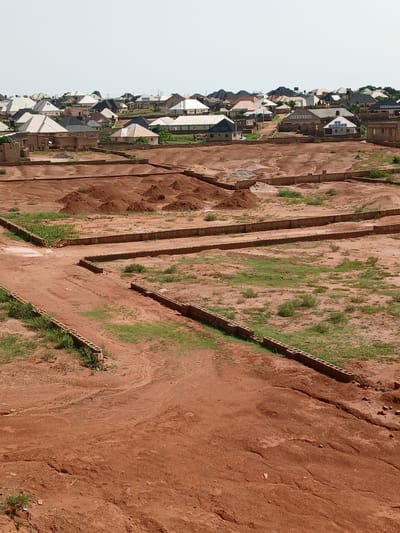 The image depicts a residential development in Katon Rikkos, Nigeria. The scene shows a landscape with many houses of varying sizes and architectural styles, characterized by their distinct roofing materials and colors, primarily white and dark grey. The houses are situated on a gently sloping terrain, with some appearing recently constructed and others more established.
In the foreground, the environment is a construction site. Red earth and piles of dirt are visible, indicating ongoing development. Low brick walls, possibly foundations or boundary markers, delineate plots of land. Patches of green grass and sparse vegetation are scattered throughout the construction area, suggesting a semi-arid climate. The sky is clear and bright, indicating daytime.
No people are visible in the image. The overall impression is one of a growing suburban or semi-rural community undergoing expansion. The presence of solar panels on some rooftops suggests a degree of modernization and awareness of renewable energy in the area.