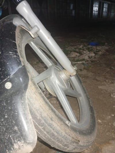 The image depicts a close-up, dimly lit view of a motorcycle's front wheel assembly in Shella ward, Kenya. The black front fender, silver-colored multi-spoke alloy wheel, and gray front fork stanchions are prominently visible. The black tire is heavily soiled with dirt and exhibits a significant deformation on its lower sidewall, consistent with a flat tire or rim damage. The ground beneath the wheel consists of uneven, unpaved dirt with scattered debris and sparse vegetation. In the dark background, indistinct building structures with faintly glowing rectangular lights are visible. A small blue object is discernible on the ground to the right of the wheel. No individuals or active interactions are present in the scene.
