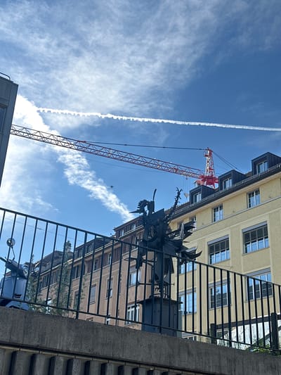 An upward-angled, outdoor photograph reveals a construction scene with a crane in Winterthur, Switzerland. The scene is partially obstructed by a black, wrought-iron fence and concrete structures in the foreground.
The sky is bright blue, streaked with wispy clouds and a prominent contrail. A red and white construction crane dominates the left side of the frame, its lattice structure clearly visible against the sky.
Buildings are visible through the gaps in the fence and next to the crane. A light yellow building with multiple rows of windows is seen. A portion of a brown building is also visible. A small dark shape, possibly a bird, is located between the crane and the fence.
The concrete structures and wrought-iron fence are located in the foreground, partially obscuring the view.
A blue scooter is partially visible behind the fence.