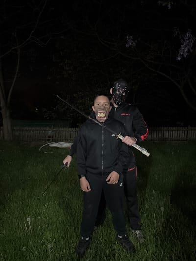 The image depicts two individuals in a grassy outdoor area, likely at night or in low light conditions. One individual is wearing a black hockey mask, and the other is wearing a mask with prominent teeth. Both are dressed in black tracksuits with red detailing that reads "ASIAR." The individual with the hockey mask is standing over the individual wearing the teeth mask, holding a decorative sword at the neck of the other individual. The masked individual on the ground is holding another sword in their left hand, blade pointed in a downward trajectory. The environment includes a dark tree with light purple flowers in the background, a fence with white pipes behind the person on the ground, and grass covering the surface of the ground. The location, Goritsa, Bulgaria is not confirmed.