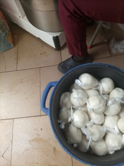 In Opete, Nigeria, an indoor scene displays a dark blue plastic bucket in the foreground, filled with approximately two dozen spherical, off-white food portions, each individually wrapped in clear plastic and secured with a knot. To the upper right, the lower portion of a person's legs is visible, clad in maroon trousers and dark open-backed clogs. This individual is positioned near a large, white industrial-style machine with a metallic bowl, visible in the upper left background. The machine rests on a white base with black protective feet. A metal stool leg with a red cap is partially visible behind the person's legs. The floor is tiled with light-colored, possibly beige, square tiles, exhibiting minor stains and grout lines. A patterned bag is partially visible on the far left.