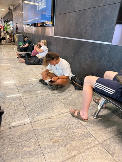 The image depicts an indoor waiting area, possibly at an airport or train station, in Pendik, Turkey. Several individuals are seen sitting on the floor and a bench. In the foreground, a young man with long, curly hair is sitting cross-legged on the tiled floor, engrossed in reading a book. He is wearing a white t-shirt and brown shorts, with several bracelets on his wrists. To his left, a woman with blonde hair pulled back is sitting with her legs crossed, looking at her phone. Another woman, with dark hair, is seated next to her, also looking at her phone. Behind them, a man wearing a dark t-shirt and a black cap is seated, facing away from the camera. There are several pieces of luggage scattered around the individuals. Further in the background, other people can be seen, some standing and some sitting. The environment appears to be a modern interior with a large screen displaying text in Turkish on the wall. The floor is made of large, light-colored tiles with a subtle patterned texture. A bench with chrome legs is visible in the right side of the frame.