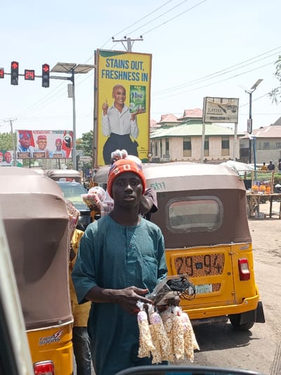The image depicts an outdoor street scene in Hotoro, Nigeria, under daylight conditions. In the immediate foreground, a young male individual with dark skin, wearing an orange and white patterned knitted beanie and a teal button-down tunic, stands looking directly forward. He holds several small clear plastic bags filled with a white, presumably food-related, substance in his left hand, and a larger clear plastic bag containing black cables in his right hand.
He is positioned between two yellow and brown tricycle taxis. The left taxi displays "TWIN SPARK" branding. The right taxi has a partially visible Nigerian license plate with "PG QX" on the top line, and a white sticker with "S11" on its brown canopy.
In the mid-ground, a prominent vertical yellow billboard features a smiling woman holding a "Viva" brand detergent product, with the slogan "STAINS OUT, FRESHNESS IN." To the left of this billboard, another billboard displays multiple male political figures and the text "RENEWED HOPE... STRONGER NIGERIA." Further left, a pole supports traffic lights, with a digital red "24" countdown visible.
The background includes various buildings, notably one with a green roof behind the Viva billboard. A "JUPITER" sign and a solar-powered street light pole are also present. Power lines are visible spanning across the sky. In the far background, additional people and what appear to be market stalls or vendor setups are discernible. The ground consists of both paved and unpaved surfaces.