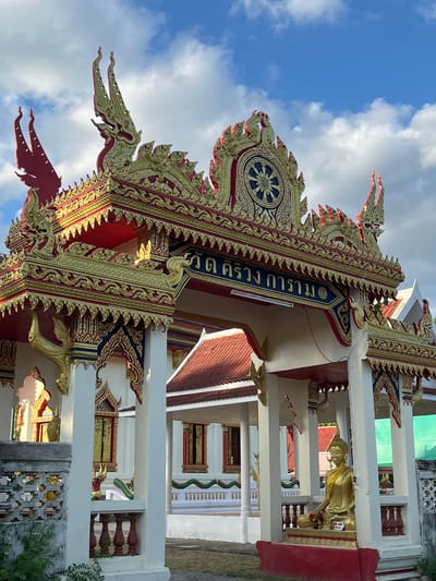 The image shows the ornate entrance to a Buddhist temple in Baan Taling Ngam, Thailand. The entrance features a multi-tiered, gold-leafed roof adorned with intricate carvings and mythical serpent-like figures (naga). A large Dharma wheel symbol is prominently displayed at the apex of the archway. Below the arch, in a small shrine, sits a golden statue of Buddha. The temple buildings in the background have traditional red tiled roofs and decorative windows. The scene is set against a bright blue sky with scattered white clouds, suggesting it is daytime. The overall impression is one of cultural and religious significance, with a focus on the detailed architecture and symbolic imagery characteristic of Thai Buddhist temples.