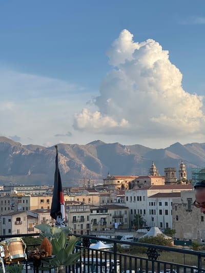 This is a vertical, eye-level shot of a cityscape in Palermo, Italy, with mountains in the background. A person's head and shoulders are visible in the top right corner, wearing sunglasses and looking down. In the foreground, a black flag with white and red designs is partially visible, along with a potted plant and what appears to be a drum.
The scene looks out over a dense collection of buildings with terracotta roofs and light-colored walls, typical of Mediterranean architecture. Some buildings have visible air conditioning units. In the mid-ground, several church domes and bell towers rise above the rooftops, indicating a historic urban center. The mountains behind the city are rugged and dry, with some visible paths or roads winding up their slopes. A large, fluffy white cloud dominates the upper left portion of the sky, contrasting with the clear blue sky in other areas.
The lighting suggests it is daytime, likely late afternoon given the warm tones in the sky and on the buildings. The weather appears clear and sunny, with the cloud adding a dramatic element to the sky. The presence of the flag and the drum might suggest a festive occasion or a gathering. There is no discernible text visible in the image.