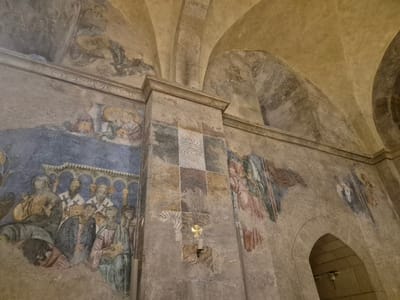 The image shows the interior of a church with fresco paintings on its walls and arched ceiling. The frescoes depict religious figures, including what appear to be saints or religious leaders, some with halos, and possibly angels. The paintings are in a somewhat faded, ancient style.
On the left wall, a group of figures with beards and religious attire are depicted, standing behind an arched structure. One figure in the foreground is seated. Above this scene, on the ceiling, there are more faded paintings.
A central column divides the wall. The column itself is decorated with geometric patterns and some of the same plasterwork as the walls. On this column, there is a gold cross mounted above a lit candle.
To the right of the column, an angel with large wings is painted, with another figure, possibly a saint, to its right. Further to the right, an arched doorway leads to a darker space. The overall environment suggests a historical religious site.
This scene is located in the Crusader period church, known as the Church of the Annunciation or St. Mary's Abbey, in Abu Gosh, Israel.