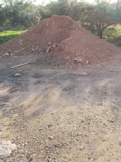 The scene is located in Lo Ferro, Spain. The image presents an outdoor, unpaved area. A large, conical mound of reddish-brown soil mixed with rocks occupies the central and mid-ground. The foreground consists of compacted light brown and gray dirt with scattered gravel and small stones. A light-colored, cylindrical wooden handle, approximately 0.5 meters long, lies horizontally on the ground to the left of the soil mound. Darker patches, consistent with ash or disturbed earth, are visible on the ground near the handle. Multiple long, distinct shadows stretch across the foreground from right to left, originating from unseen objects. In the background, behind the soil mound, a dense line of mature olive trees with green foliage and visible dark trunks extends across the width of the frame. A patch of green grass is discernible on the far left, adjacent to the base of the soil mound. No individuals, actions, or interactions are present.
