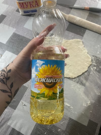 The image shows a person's hand holding a bottle of "Нежинское" (Nezhinskoe) sunflower oil. The hand has long, light-colored artificial nails and a tattoo of leaves on the wrist and forearm. The bottle of sunflower oil is clear plastic with a blue label featuring a large sunflower and fields of sunflowers. Text on the label reads "подсолнечное масло" (sunflower oil), "Нежинское" (Nezhinskoe), and "Высший сорт" (Highest grade), as well as "РАФИНИРОВАННОЕ ДЕЗОДОРИРОВАННОЕ ВЫМОРОЖЕННОЕ" (refined, deodorized, and winterized).
The setting appears to be a kitchen or dining area, with a grey and white checkered tablecloth visible. In the background, there is a rolling pin, a partially rolled-out piece of dough, and a knife. A package labeled "МУКА" (flour) is also partially visible on the left side of the image. The scene suggests the preparation of food, likely baking or cooking. There are no specific cues about the time of day or weather. The overall mood is neutral, focusing on the task at hand.