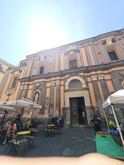 This is a daytime, low-angle, vertical, outdoor shot of the facade of the Church of Gesù Nuovo in Naples, Italy. The church is a Baroque-style building with a peach-colored facade featuring gray pilasters and decorative elements. The main entrance is a large wooden door with ornate carvings, flanked by a black wrought-iron gate. Above the entrance, a semicircular arch is visible, and further up, there are windows and sculptural details. The building is adorned with graffiti, particularly near the ground level. In front of the church, there is a public square paved with dark cobblestones. Several white umbrellas with "O'Café" branding are set up, suggesting outdoor seating for a cafe. Multiple small, yellow and dark-colored tables and chairs are arranged in the square. A few people are seated at the tables, engaged in conversation or looking at their phones. A man in a black t-shirt and blue jeans is standing near a green-covered table to the right of the entrance, with some items displayed on the table. The sky is bright blue with visible lens flare from the sun in the upper left corner. There is a statue of a religious figure on the facade of the church, to the left of the main entrance. A bicycle is parked near the church entrance.