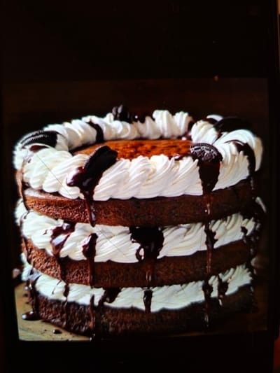 This is a close-up, low-angle, indoor shot of a three-layer chocolate cake adorned with white frosting and drizzles of chocolate sauce. Several Oreo cookies are placed on top and between the layers. The cake is positioned on a wooden surface against a dark background. The image is slightly blurry and has some glare. There is no indication of the city of Medvezhyi Ozyora, Russia, or any people, actions, or interactions.