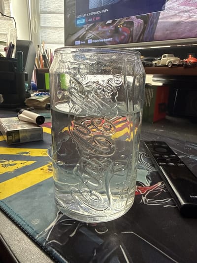 The image depicts a close-up, eye-level shot of a glass of clear liquid, likely water, with embossed branding that resembles a soda bottle, placed on a desk. To the left of the glass is a pack of cigarettes and a lighter. In the background, a computer monitor displays text in Cyrillic, possibly related to a game or software. A few small toy cars are visible on a shelf above the desk. The desk itself is covered by a mousepad with a graphic design. The scene appears to be indoors, in what could be a home office or personal workspace. There is no indication of the city of Grătiești, Moldova.