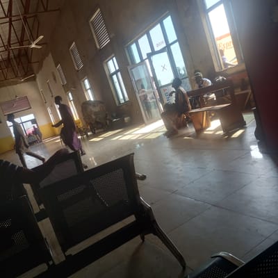 The image shows the interior of a waiting area, likely a bus station or similar transport hub, in Hayin Banki, Nigeria. The space has a tiled floor and concrete walls, with large windows providing natural light. Several ceiling fans are visible, indicating a warm climate. In the foreground, there are dark metal benches with perforated seats.
Several individuals are present in the scene. In the mid-ground, a person in a purple garment walks towards the left side of the frame. To their right, a person wearing a hat and white shirt sits on a bench, with a bag next to them. Further in the background, a person in a brown garment sits on a bench near a window. Through the windows, a truck or vehicle with the word "Express" visible is partially seen.
Other objects in the scene include a cart with a large bundle covered in a green tarp, and various bags and luggage. The overall impression is of a functional, somewhat rustic waiting area. The lighting suggests it is daytime, with strong shadows cast by the windows. The architectural style and the presence of the truck with "Express" branding suggest this is a commercial transport hub.