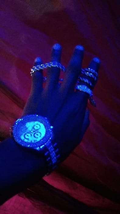 A human left hand, adorned with multiple luminous jewelry pieces, is presented under blue/ultraviolet light against a dark red draped fabric background in Ndeiya, Kenya. The hand is viewed from the dorsal side with fingers slightly splayed and pointing upwards. A large, circular wristwatch is worn on the wrist, featuring an encrusted bezel and three luminous chronograph sub-dials on its face; the watch's metal band is also encrusted and glowing. Rings, all appearing encrusted with small, glowing elements, are visible on the index, middle, and ring fingers. The fingernails of the visible fingers also exhibit a subtle glow. No other individuals or specific environmental markers are discernible beyond the background fabric.
