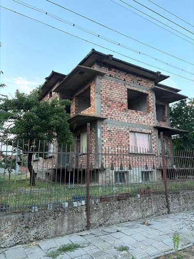 The image depicts an unfinished, two-story brick building under construction in Dolni Dabnik, Bulgaria. The building's exterior is exposed brickwork with concrete horizontal slabs serving as floors or balconies. Several window openings are visible, some appearing as empty frames. A mature tree with green foliage stands to the left of the building, partially obscuring the structure. The property is enclosed by a rusty metal fence with pointed finials. The foreground shows a concrete sidewalk with some cracks and sparse vegetation. The sky is clear and light blue, with power lines running horizontally across the upper portion of the frame. The overall scene suggests a stalled construction project in an outdoor, residential-like setting.