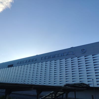 This image depicts the exterior of Arlanda Terminal 5, located in Stockholm-Arlanda, Sweden. The prominent feature is the upper section of a modern building, identifiable by the blue-outlined signage "ARLANDA TERMINAL" and a circled number "5" on its facade. The building's upper exterior is composed of a light grey material, beneath which a section of the wall features numerous horizontally aligned, textured rectangular panels arranged in a grid pattern. These panels create a series of triangular or wedge-shaped shadows, adding depth to the facade. Below the building, an elevated concrete structure, possibly a bridge or platform, is visible, with support pillars extending downwards. Further below, faint lines suggest what could be railway tracks or a road. A tall, slender lamppost is present on the right side of the lower frame. The sky occupies the upper portion of the image, displaying a clear, bright blue color, with a fragment of a white cloud visible in the upper left corner. The lighting appears uniform, suggesting a daytime scene without harsh shadows. No individuals or active elements are discernible within the frame.