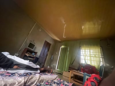The image depicts a cluttered room with two beds in the foreground, one covered with a floral-patterned sheet and the other with a white and grey blanket. A person is lying on the bed covered with the white and grey blanket, wearing black and white clothing. The room has a wooden ceiling and plastered walls. To the left, there is a wooden shelf unit with a television on top. A brown wooden door is visible on the left side of the room, and a camera tripod stands near it. On the right side, there's a window covered with light green curtains, and beyond that, another window with yellow curtains, letting in natural light. A wooden table or bench is in front of the window, with a red bag and a red object on it. The overall impression is of a lived-in, somewhat disheveled space. The scene suggests a daytime setting, possibly indoors in a residential area in Aiyegunle, Nigeria. There is no visible text in the image.