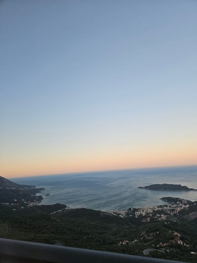The image captures a panoramic view of Brajici, Montenegro, at dusk. The sky transitions from a pale blue at the top to a soft gradient of peach and pink near the horizon, meeting the calm, blue sea. Below, the coastline is dotted with buildings, a town nestled amongst lush green hills. The water is a deep blue, with gentle ripples reflecting the fading light. To the right, a small, dark island rises from the sea. In the foreground, a dark, railing-like object occupies the bottom of the frame, suggesting the viewpoint is from an elevated position, possibly a balcony or a lookout point. The overall scene is serene and tranquil, showcasing the natural beauty of the Montenegrin coast.