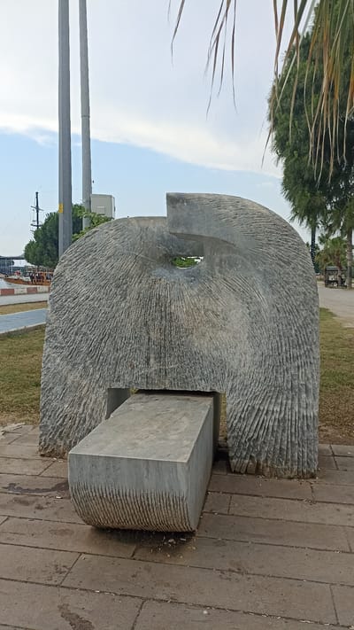 A concrete sculpture resembling a stylized ear or a large shell stands in a park setting. The sculpture is massive, with a rough, textured surface, and features a central opening. A rectangular concrete bench is placed in front of the sculpture, also with a textured surface. The sculpture and bench are situated on a paved walkway made of large stone tiles.

Behind the sculpture, there is a grassy area with scattered trees, including palm trees and a larger evergreen. In the distance, a tall grey pole with some equipment attached is visible, along with a hint of a body of water and a boat. The sky is partly cloudy, suggesting daytime. The overall scene appears calm and public, possibly a park or a public art installation. There is no visible text in the image.