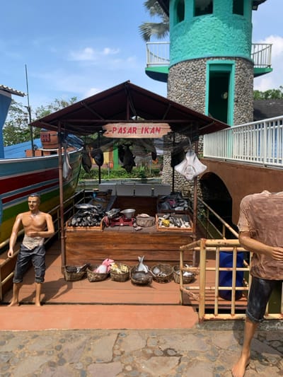 The image shows a fish market stall in the city of Batu, Indonesia, located at latitude -7.883 and longitude 112.526. The stall is made of wood and has a roof. It is located next to a small, wooden boat.  The stall is decorated with a sign that says "PASAR IKAN" in red letters on a white background.  It has a variety of fish on display, including stingrays, catfish, and tuna. There are baskets of fish on the counter and some hanging from the roof. A man in a blue t-shirt stands next to a railing.  The stall is located next to a green building with a stone facade and a wooden railing. There is a blue barrel nearby. The ground is paved with cobbles and the sky is clear.