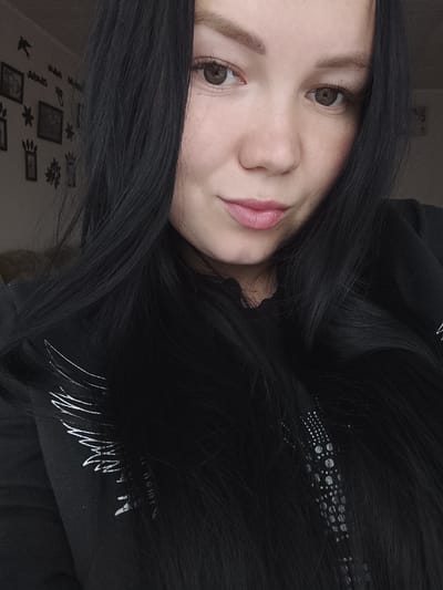 A close-up, medium-lit shot features a woman with long, straight black hair framing her face. Her skin is fair, with visible freckles around her nose. She has brown eyes, light eyebrows, and pink lips. She's wearing a black hooded sweatshirt, with a white wing design and a rhinestone detail visible on the front. The background is a lightly colored wall decorated with framed pictures and silhouette cutouts. There is no evidence to indicate that the location is Kuyeda, Russia.