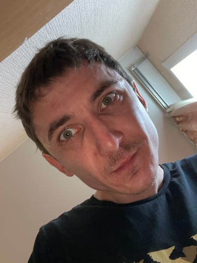 A close-up, low-angle selfie shows a man with short, dark hair and light green eyes. He is wearing a black t-shirt. The background appears to be an indoor setting with a textured wall and a window with blinds. The lighting suggests it is daytime. The man's expression is curious or surprised, with his mouth slightly open and eyebrows raised. The overall impression is a candid, unposed shot. The location is Laishevo, Russia.