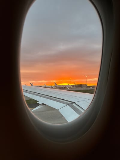 The image shows a view from an airplane window. Visible through the window is a vibrant orange sky, likely during sunrise or sunset. An airplane wing is also visible on the right side of the window. Beyond the wing, the ground contains airport infrastructure, including buildings and lampposts. Based on location, the scene likely captures an image taken while on a plane at Charles De Gaulle airport located near Le Mesnil-Amelot, France.