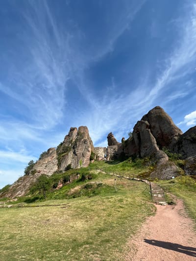 This is a wide-angle, ground-level shot of the Belogradchik Rocks in Belogradchik, Bulgaria, under a bright blue sky with wispy clouds. In the foreground, a reddish-brown dirt path curves to the right, leading towards a set of stone stairs. The stairs ascend a grassy slope that is dotted with green vegetation. To the left of the stairs, a wooden fence with posts borders the path.
Further up the slope, imposing sandstone rock formations rise dramatically, their surfaces a mixture of reddish-brown and grey tones, with patches of green vegetation clinging to their sides. The rocks are irregularly shaped and appear weathered. On one of the higher rock formations, the stone ruins of a fortress are visible, with arched doorways and walls integrated into the natural rock. A few small figures of people can be seen near the fortress ruins, standing on the stairs or pathways. The overall environment is natural and rugged, with a sense of historical significance conveyed by the ancient fortress.