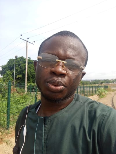 A close-up, upward-angled selfie features a man of African descent. He is positioned slightly off-center to the left, looking directly at the camera with a neutral expression and slightly pursed lips. He wears gold-rimmed glasses and a dark green garment, possibly a traditional tunic, with a distinctive light green stitched pattern around the placket. White wired earbuds are visible, with one wire hanging down his chest and the earpiece resting on his shoulder. He has short dark hair and a sparse beard or stubble.
The background reveals an outdoor setting under a bright, slightly hazy sky. To the man's left, a tall utility pole with several wires stretches upwards, with dense green trees and a street light further in the distance. A green metal fence runs horizontally behind him, separating the foreground from a more open, possibly paved area or road on the right. In the far background, distant buildings or structures are faintly visible under the wide expanse of the sky. The overall scene suggests a bright, sunny day in what appears to be an urban or semi-urban environment in Kuchingoro, Nigeria. No visible text is present in the image.