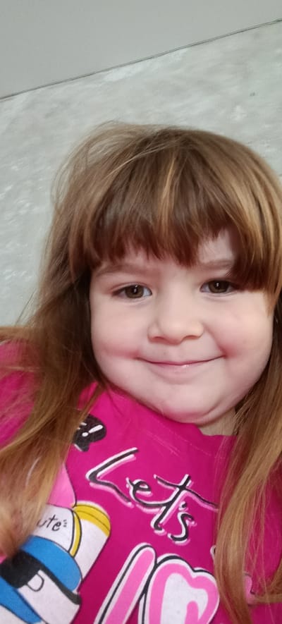 A young girl with brown hair and brown eyes is captured in a close-up, selfie-style portrait. She is wearing a bright pink t-shirt with a graphic design. The t-shirt features the word "Let's" in a stylized font, along with other cartoon-like graphics. Her hair is long and appears to be styled with bangs. The background is a neutral, textured gray wall. The lighting is soft and even, illuminating her face. The image is taken from a slightly upward angle. There is no indication of the specific location within Хохлы, Russia.