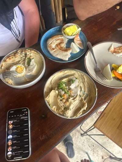 A wooden table displays several plates of Middle Eastern food. A white plate has a small portion of cooked vegetables and pita. A blue plate has pickles, onions, and pita. Two metal bowls contain hummus, one garnished with an egg. A phone is on the table displaying a list of names under the word "Affiliate." Legs and feet are visible on both sides of the table, along with a sandal.