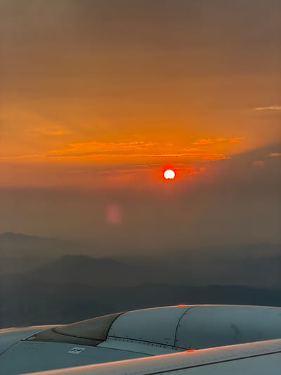 The image displays a striking aerial view from an airplane window, oriented vertically. A vibrant red sun is the focal point, positioned towards the center, appearing as a distinct orb through a heavily hazy sky. Bright orange, streaky clouds are illuminated by the sun, appearing as vertical patterns to its left. The sky's colors range from warm orange and brown near the sun and what appears to be a horizon line, to a muted gray-brown in the upper and right sections, indicating significant atmospheric haze or smoke.
A section of the airplane's wing and engine nacelle occupies the right side of the frame, reflecting the sun's reddish-orange glow along its edges. Details like rivets and panel lines are visible on the aircraft structure. Below the perceived horizon line in the lower right, distant landforms or mountains are dimly visible through the thick haze as dark, undefined shapes.
The scene captures either a sunrise or sunset, with the sun's low position and intense red hue likely amplified by atmospheric conditions such as dust or smoke. While no specific geographical markers are visible in the image, the context provided is Baiyun District, China. Faint, unreadable white text or symbols are present on the airplane's engine nacelle. The image conveys an ethereal and dramatic sense of atmospheric depth.